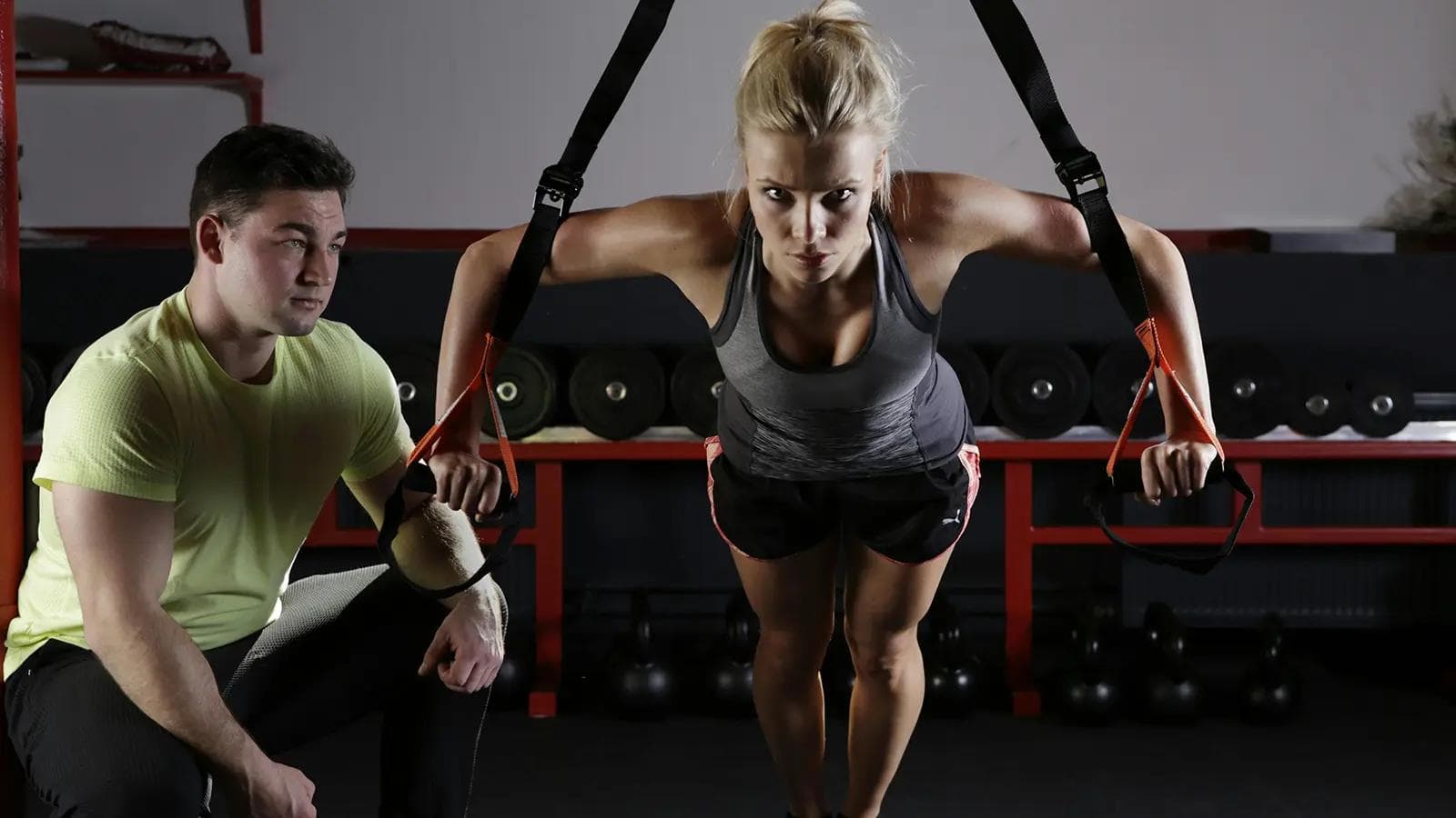 Beat 'Gymtimidation': A Beginner's Guide to Overcoming Gym Anxiety and Building Confidence Woman performing a focused suspension strap push-up in a gym while a male trainer observes her form.